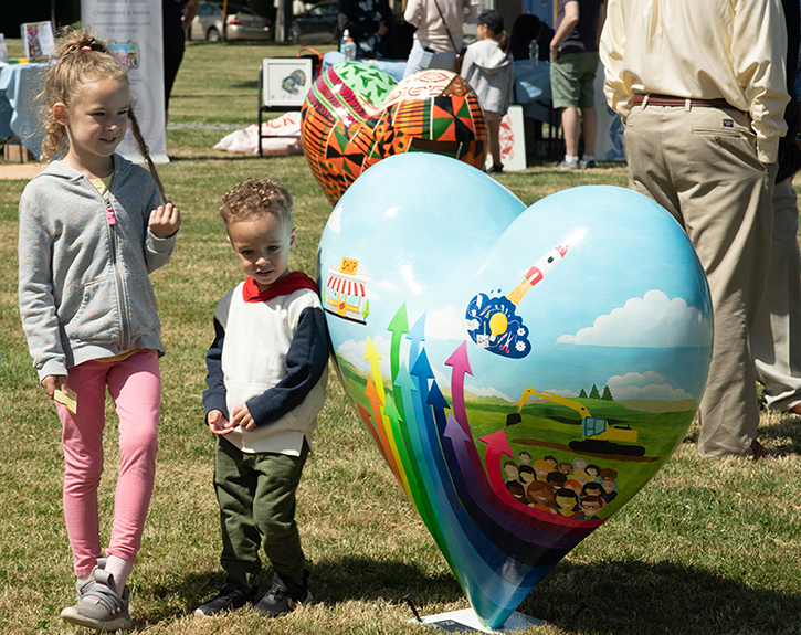 photo of 2 children standing next to a heart-shaped piece of public art with a rainbow of arrows pointing up painted on it