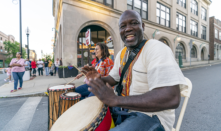 photo of a man on a side walk during a street festival drumming and smiling into the camera in the foreground. Another drummer is playing with him in the background.