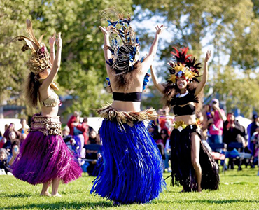 photo of an outdoor Indigenous Polynesian dance - three dancers stand on grass with their hands in the air, the sun shining on their big, colorful headpieces and full skirts